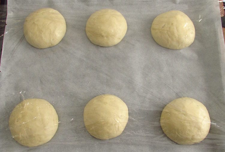 Massa de pão de alho dividida em seis num tabuleiro forrado com papel vegetal e coberta com película aderente