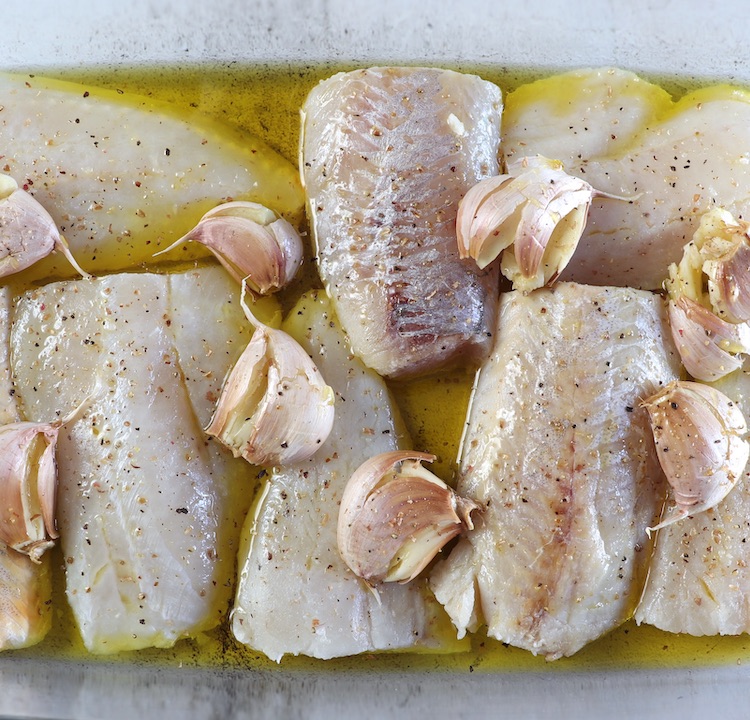 Filets de merlu assaisonnés de sel, poivre, huile d'olive et ail écrasé non pelé sur un plat de cuisson en verre