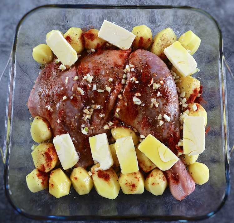 Turkey leg and peeled small potatoes seasoned with salt, pepper, nutmeg, paprika, chopped garlic, butter pieces, honey and olive oil on a glass baking dish