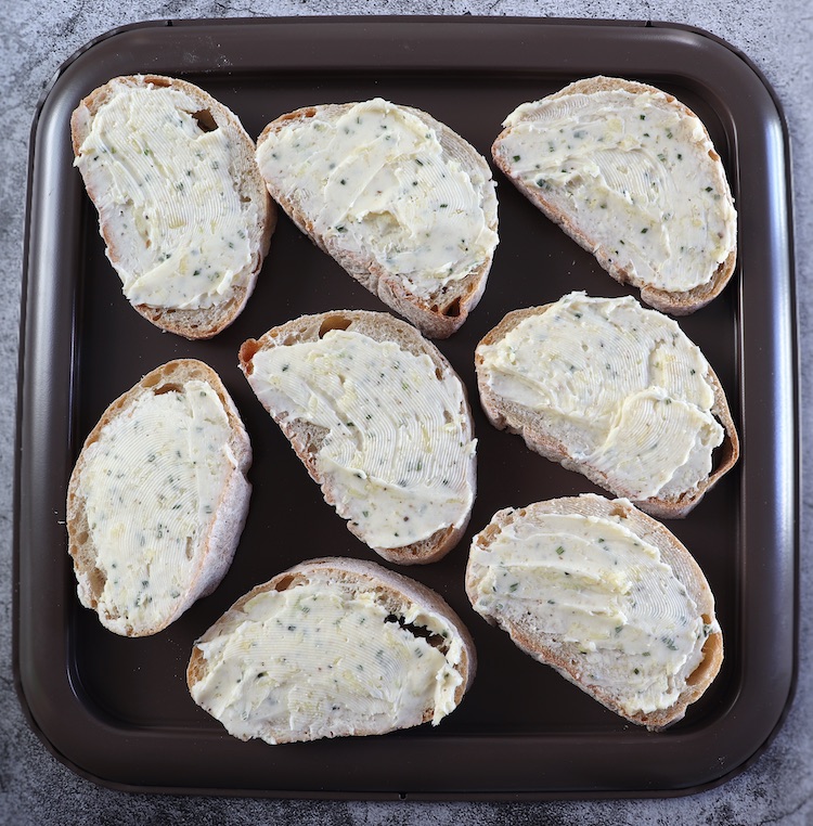 Slices of bread spread with a mixture of butter, garlic, pepper and rosemary on a baking sheet